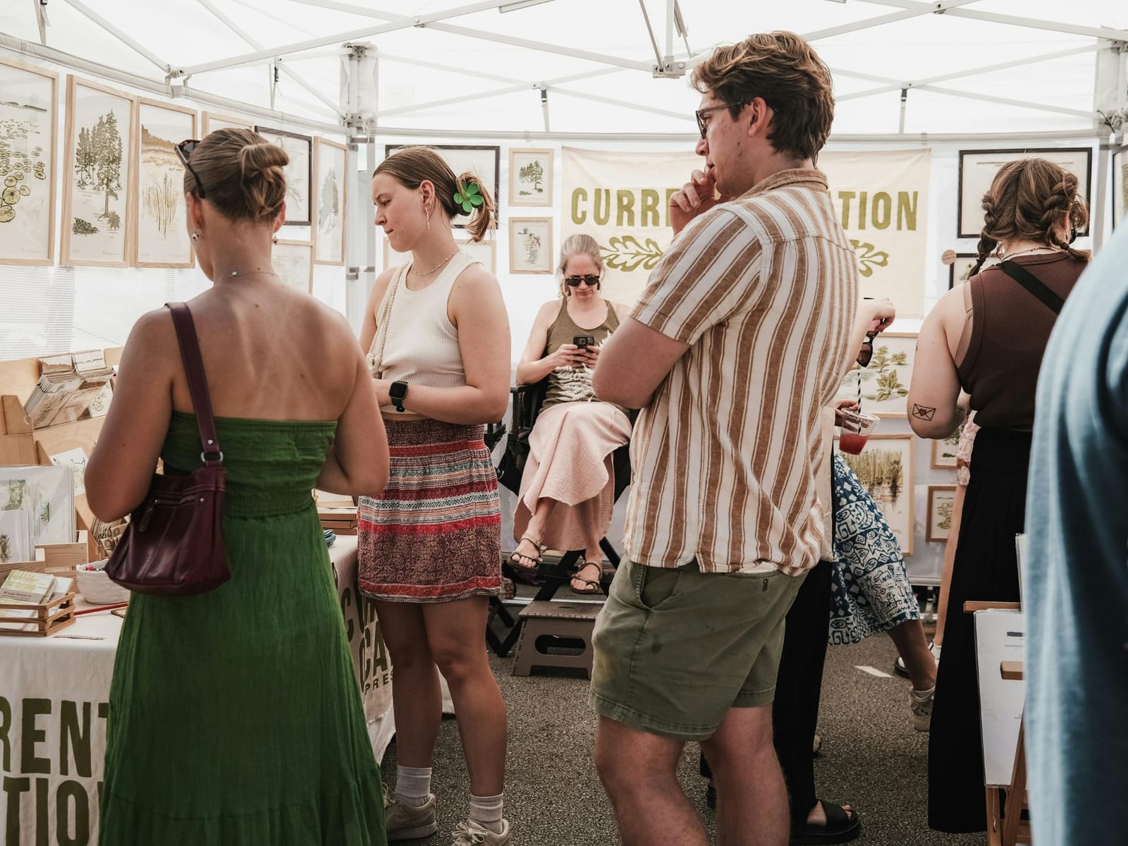 A crowded art fair booth under a white canopy, framed botanical prints and drawings hung close together on the walls while several people browse.