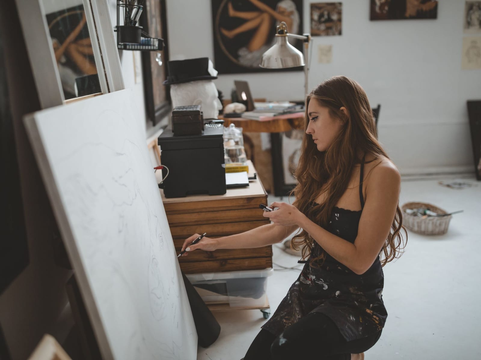 A painter sitting at an easel in her own studio, a phone in one hand and a pencil in the other, finished canvases stacked and hung behind her.