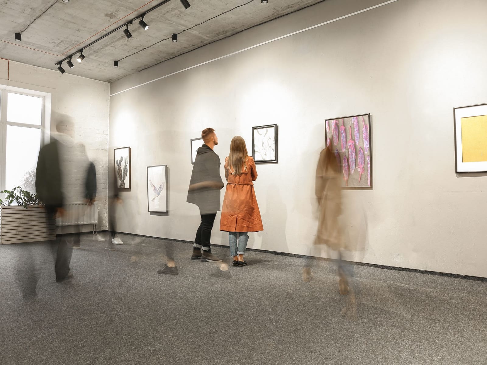 A gallery room hung with small framed works. Two people have stopped in front of one; several others are blurred, still walking.
