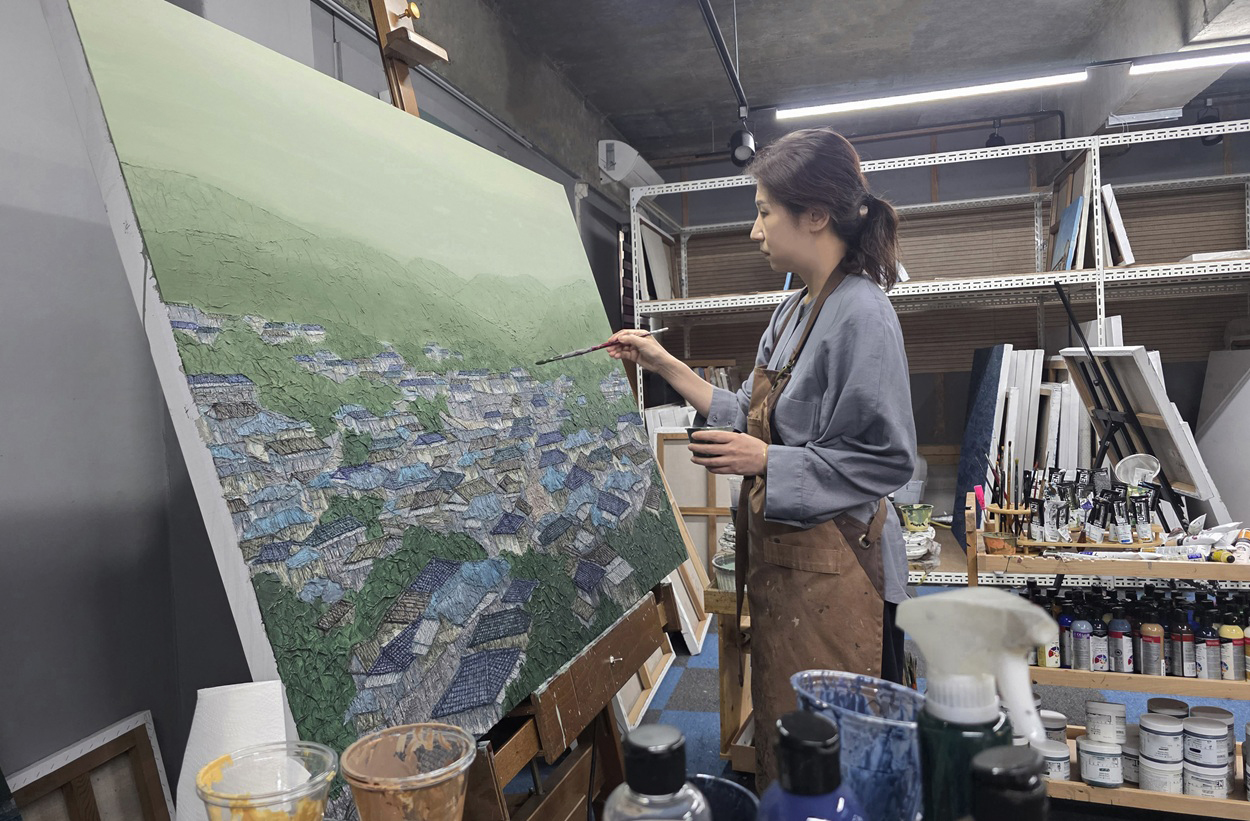 Youngju Joung painting at an easel in her studio, the canvas lit from the side so the torn paper roofs stand out in relief.