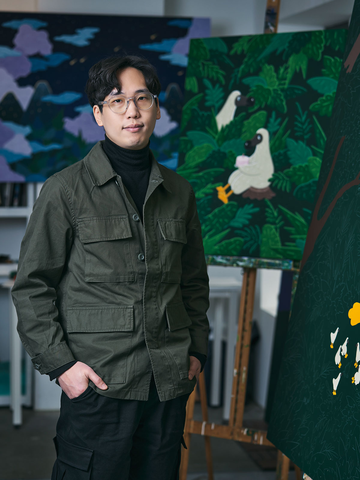 Kim Sunwoo standing in his studio in an olive jacket, with a night sky canvas behind him and a green jungle painting of two dodos on an easel.