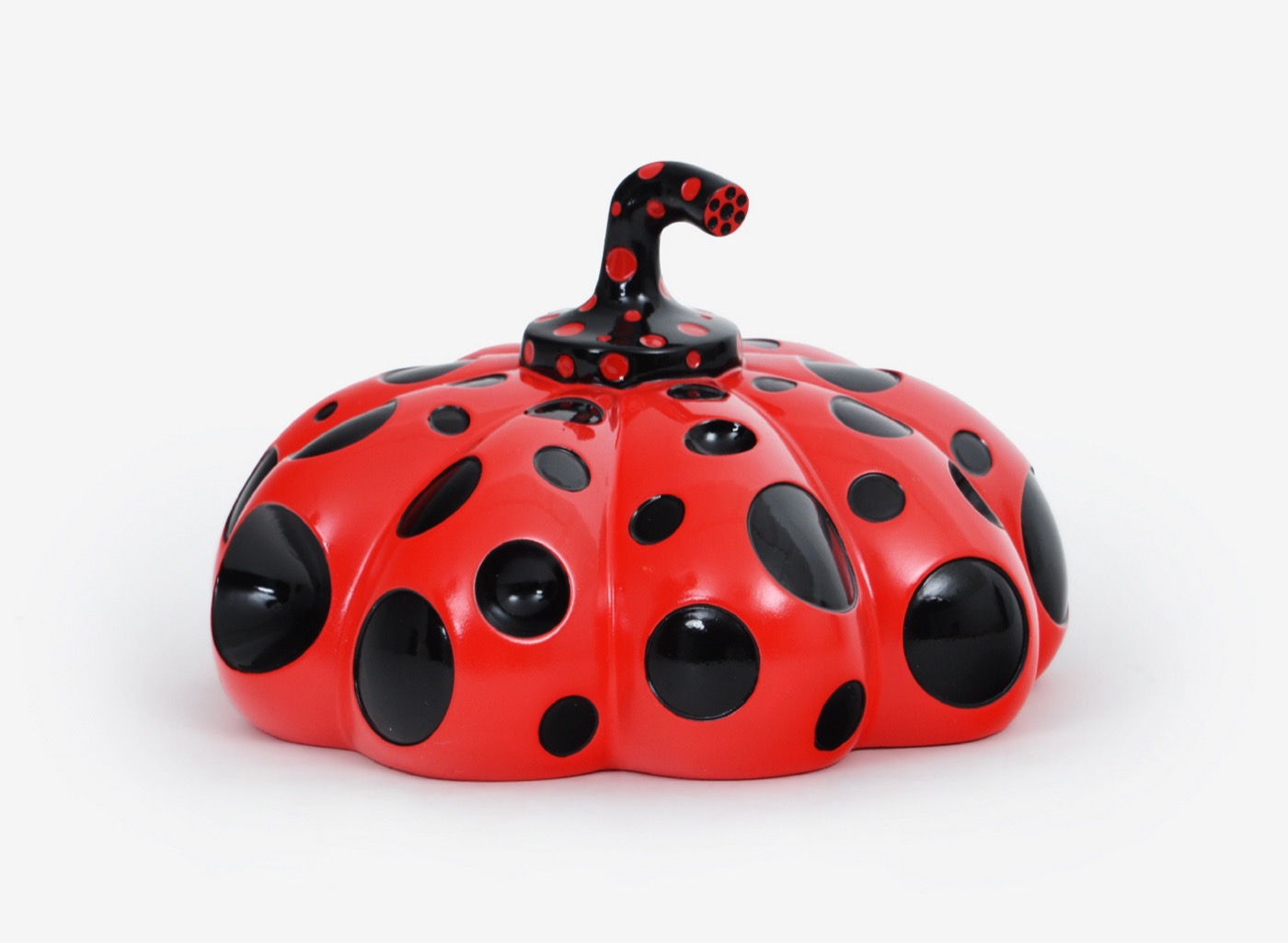 A small glossy red pumpkin with large black dots and a black spotted stem, photographed on a white background.