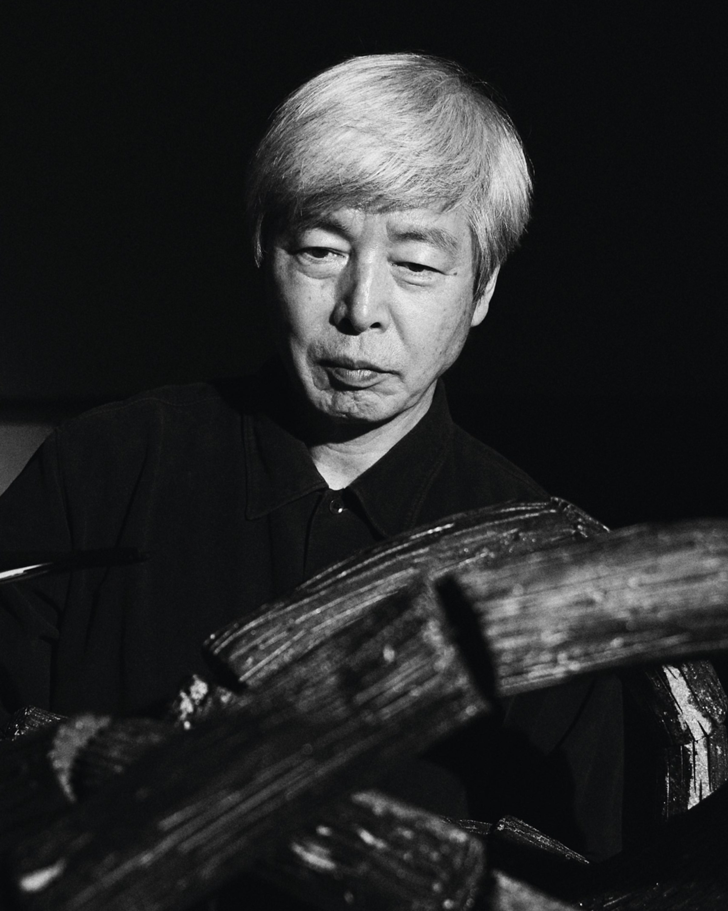 Lee Bae in black and white, looking down past a charcoal sculpture whose blackened, grooved surface fills the lower half of the frame.
