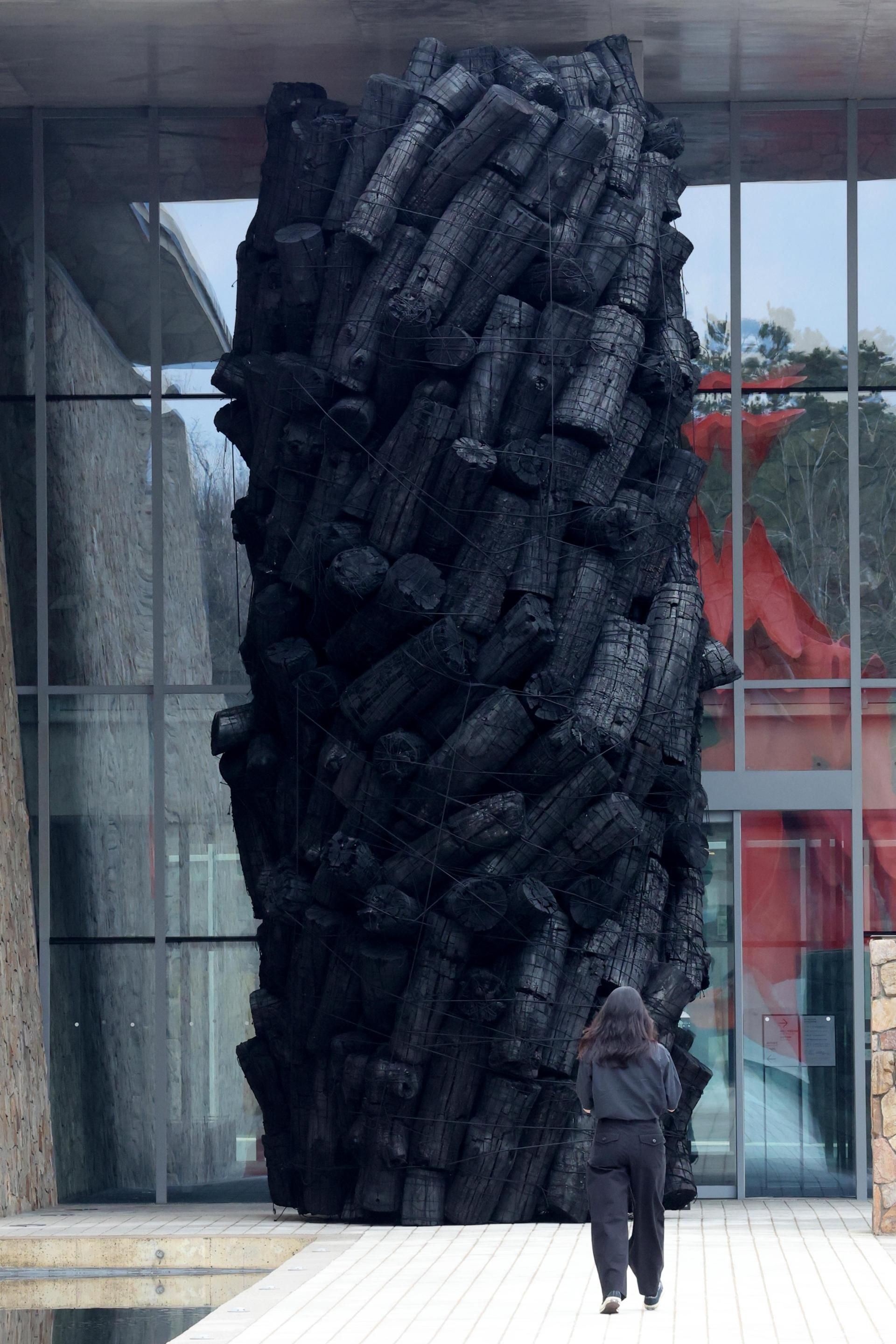 An eight metre charcoal sculpture standing at the entrance to Museum SAN, built from stacked blackened logs, with a visitor at its base for scale.