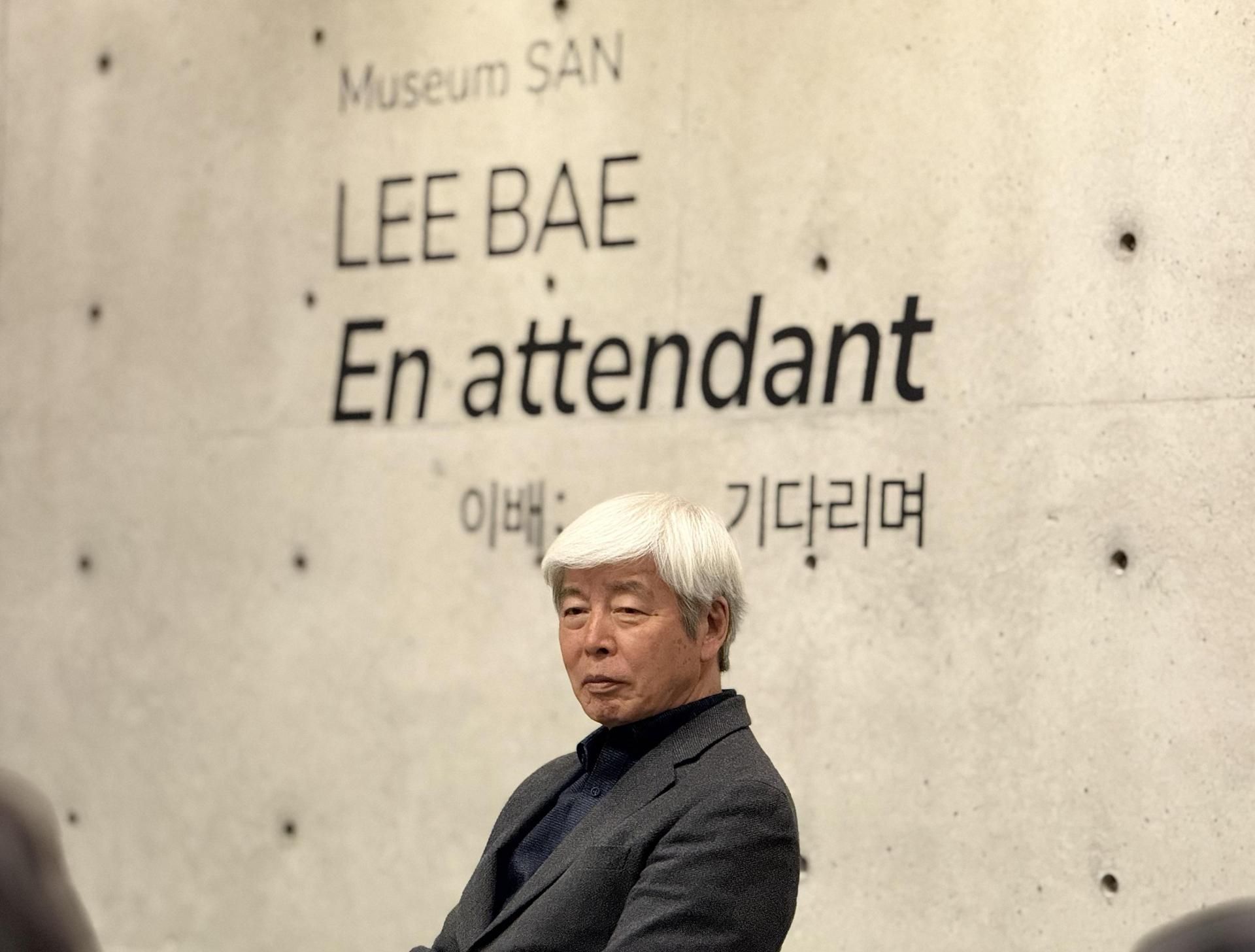 Lee Bae seated in front of a concrete wall carrying the title of his Museum SAN exhibition, En attendant, in white lettering.
