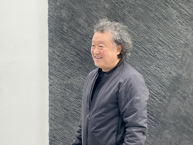 Lee Jinwoo standing and smiling in front of one of his dark charcoal works, wearing a navy jacket.
