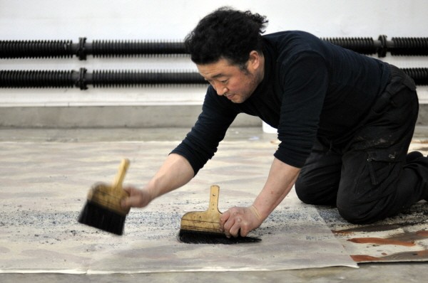 Lee Jinwoo kneeling on his studio floor, sweeping crushed charcoal across a large sheet of paper with two wide brushes.