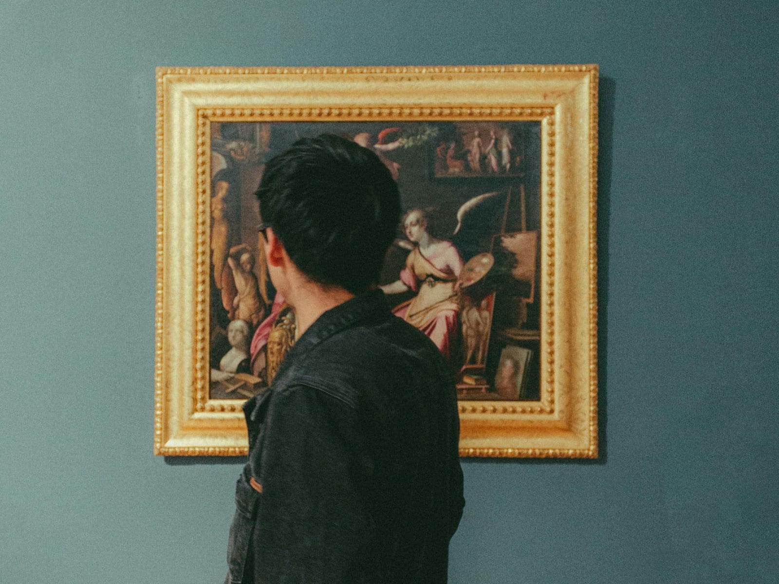 A man seen from behind, standing close to a single old master painting in a heavy gilt frame on a deep blue-green gallery wall.
