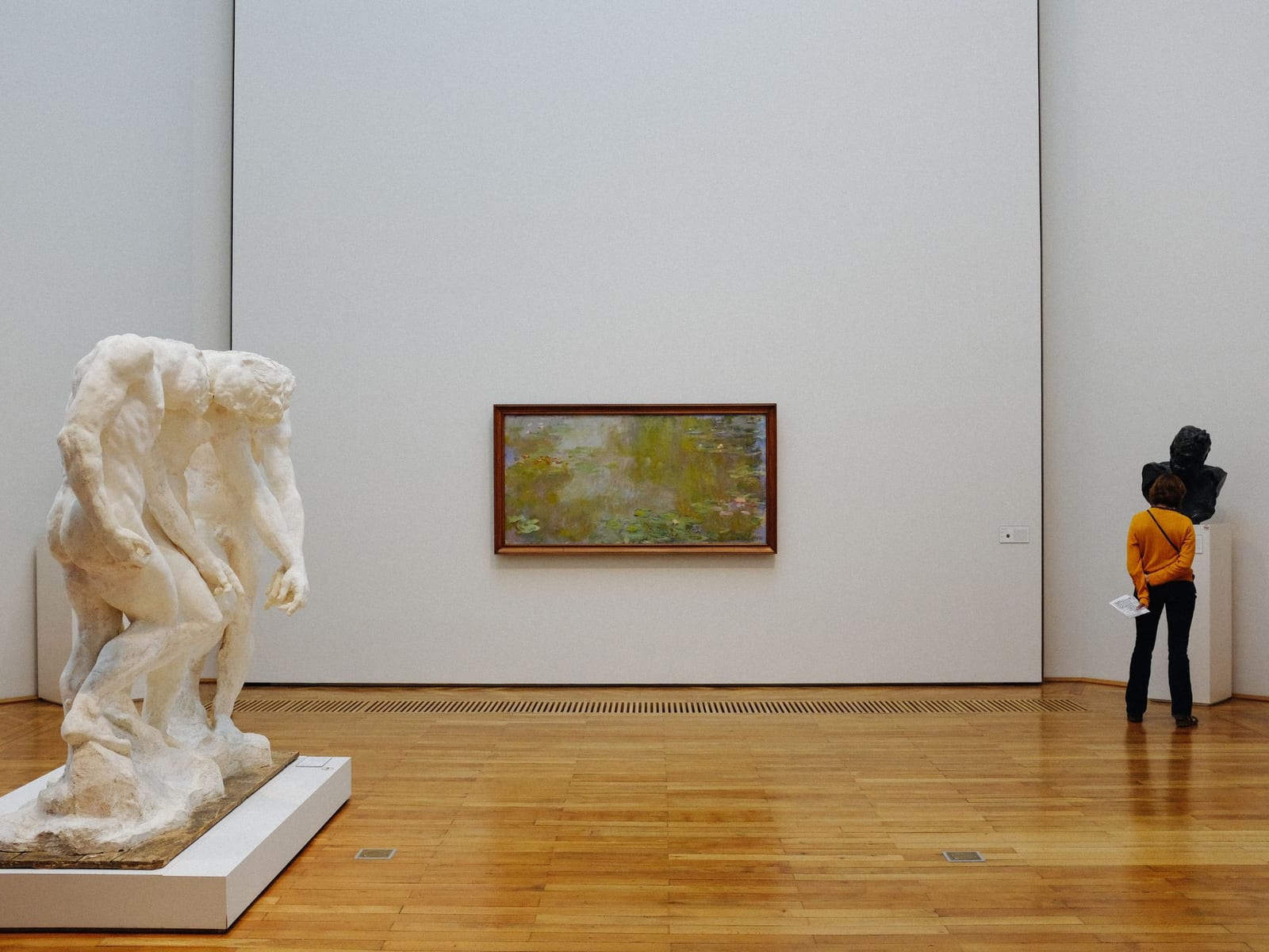 A single framed painting hung alone on a tall pale wall, with a sculpture on a plinth to one side and one visitor looking at a label.