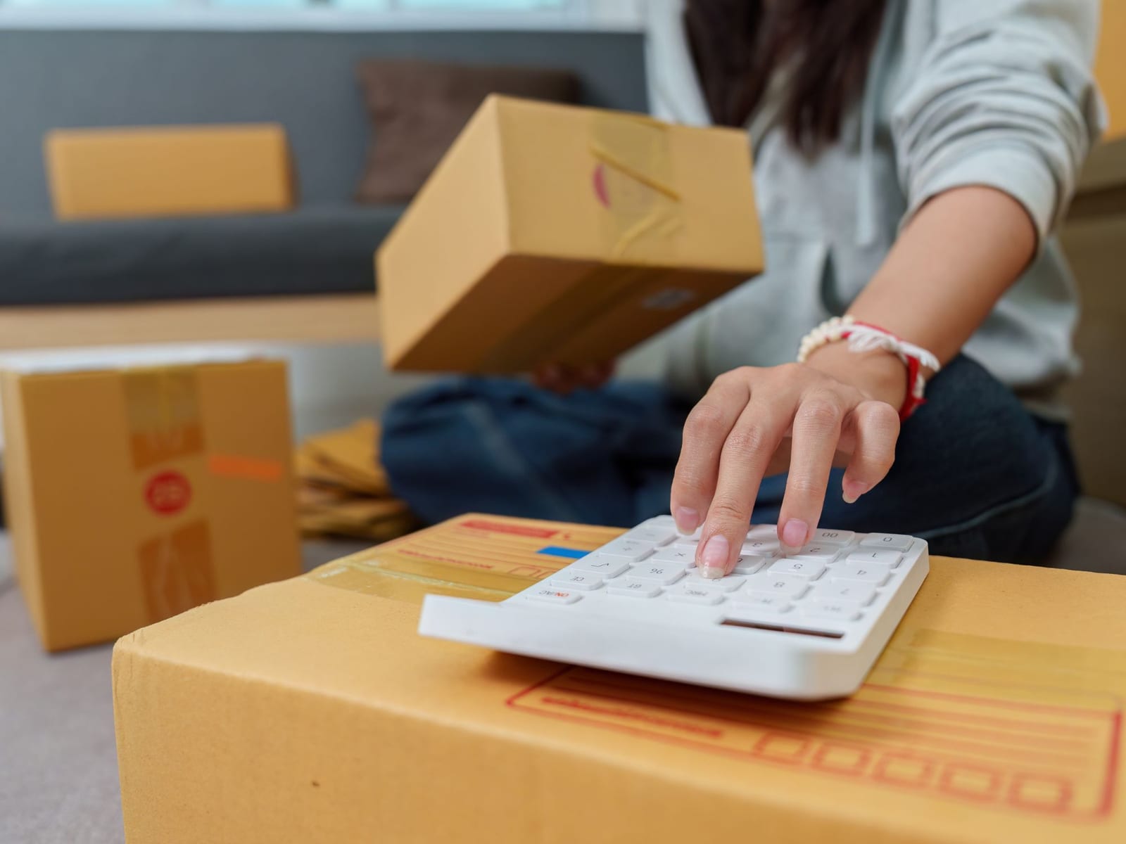 Someone kneeling on the floor beside stacked cardboard boxes, taping one shut and working out a figure on a calculator resting on the lid.